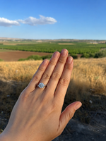 Load image into Gallery viewer, Close-up of a round solitaire natural diamond engagement ring in 18K white gold worn on hand in a scenic countryside setting.