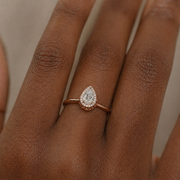 Pear-shaped diamond halo ring in rose gold with round diamonds border, worn on hand, bezel-style look