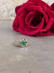 Round green emerald center ring surrounded by natural diamonds, featuring round and baguette halo stones with small princess-cut accents, displayed on a gray surface with a red flower background