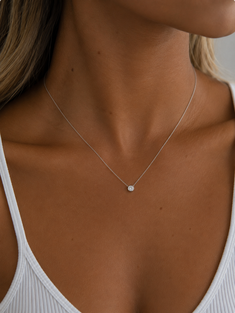 White gold necklace with a natural round diamond in a bezel setting, modeled on a woman’s neckline with a fine chain.