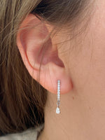 Load image into Gallery viewer, Natural diamond raindrop earrings in white gold, designed with pavé huggie hoops and dangling pear-shaped diamonds, modeled on a woman’s ear.