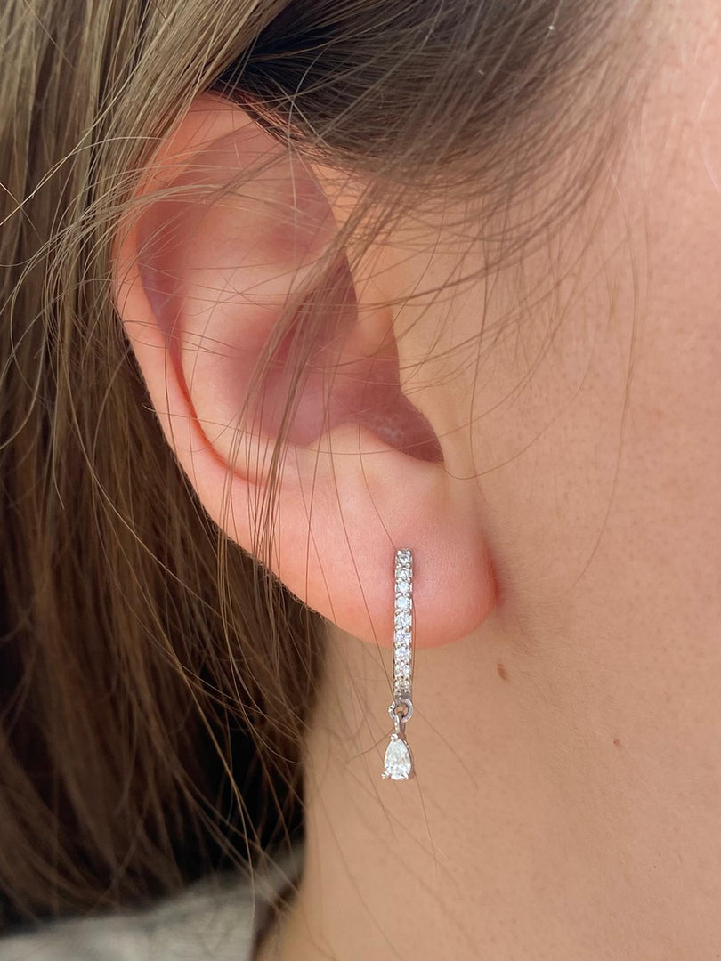 Natural diamond raindrop earrings in white gold, designed with pavé huggie hoops and dangling pear-shaped diamonds, modeled on a woman’s ear.