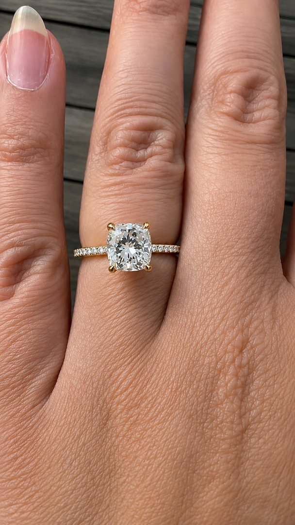 Yellow gold cushion-cut natural diamond solitaire engagement ring worn on a woman’s hand, with round pavé diamonds along the band.