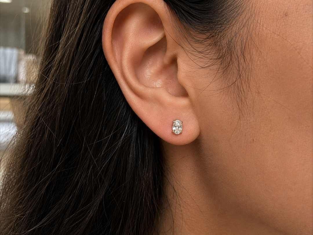 Natural oval diamond stud earrings worn on a woman’s ear with a classic four-prong solitaire setting.