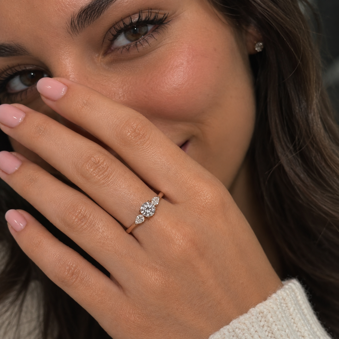 Woman wearing a lab-grown round diamond engagement ring with pear side stones in rose gold, close-up lifestyle portrait.
