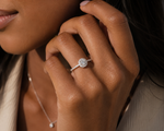Load image into Gallery viewer, White gold round halo diamond ring with side stones worn on a woman’s hand, showcasing the brilliant center diamond, halo setting, and pavé band