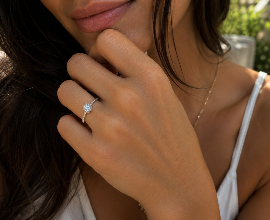 Round solitaire natural diamond ring with pavé side diamonds worn on a woman’s hand.