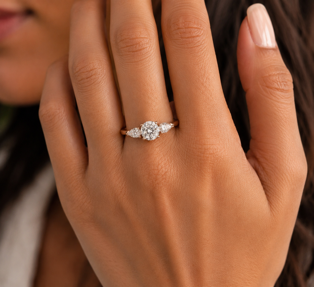 Natural diamond three-stone engagement ring in yellow gold with a round brilliant center stone and pear-shaped side diamonds, photographed on a woman’s hand.